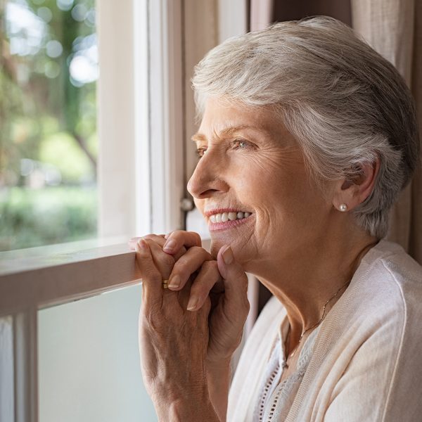 Happy senior woman standing at window and looking outside. Beautiful grandmother smiling while looking through the window. Portrait of cheerful old woman relaxing in home while standing at window sill.