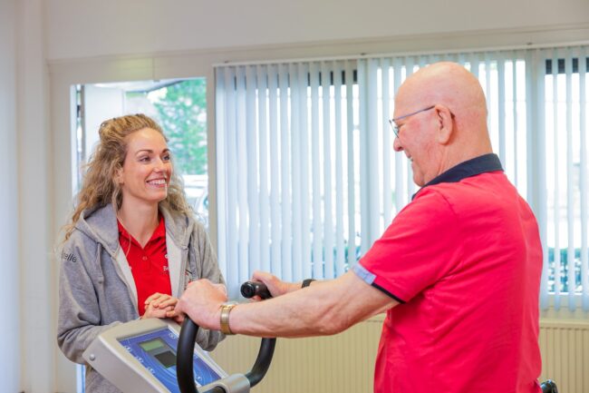 Een man staat in een kamer en heeft zijn handen vast aan een bewegingsapparaat wat helpt tegen een slijmbeursontsteking. Naast hem staat een fysiotherapeut.