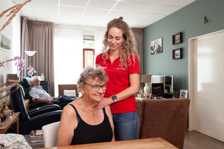 Vrouw wordt geholpen aan haar schouder na haar arm optillen met behulp van een fysiotherapeut in een rood shirt in haar huis
